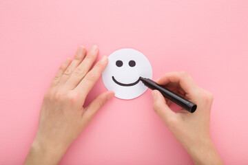 Young adult woman hand holding black marker and drawing happy smiling face on white paper on light pink table background. Pastel color. Closeup. Point of view shot. Top down view.