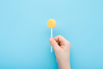 Little child fingers holding yellow lollipop on stick on light blue table background. Pastel color....