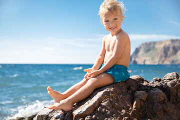 Cheerful smiling little blond boy in blue swimming trunk sitting stone against backdrop sea and sky