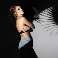 Beauty and fashion concept. Studio portrait of beautiful woman hugging herself and illuminated with bright studio light. Model wearing black bra and blue jeans. Palm leaf in dark background
