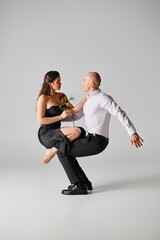 brunette woman in black dress holding red rose and balancing on laps of male dancer on grey backdrop