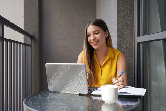 Attractive woman sits with a laptop on the balcony and works, looking intently at the screen. Beautiful girl freelancer in video conference at the notebook sitting on a balcony.