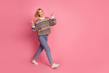 Full length photo of positive woman dressed print sweater denim pants go in empty space show v-sign isolated on pink color background