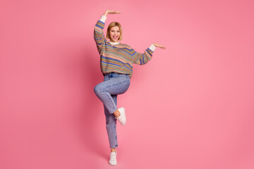 Full length photo of gorgeous woman dressed print sweater denim pants dancing raising hands up have fun isolated on pink color background