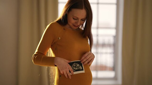 Pregnant woman looking ultrasound report. Charming pregnant female watching her ultrasound report and touching her abdomen, admiring sonography picture of her unborn baby. Expecting baby concept.