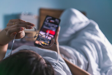 An over-the-shoulder view of a person in bed checking a smartphone with glasses in hand, signifying...