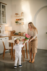 Fototapeta na wymiar Son gives mother flowers. Surprised and happy and smiling mom. The little boy goes and holds big bouquet of tulip flowers to his mother in kitchen. Child makes surprise for Mothers Day. Back view