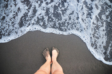 Women's feet are standing on a sandy beach. A wave of the sea or ocean during the surf. The concept of relaxation, beach and summer