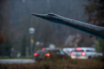 Detail Skulptur fliegende Gans mit Regentropfen vor Autoverkehr mit roten Rücklichtern in der Dämmerung
