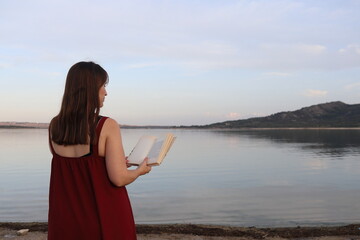 relaxed brunette haired woman in maroon dress holding a book on the bank of a river. Copy space.