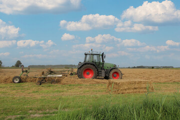 Fototapeta premium a farmer is driving with a tractor with a hay rake in a field with straw and a blue sky with clouds