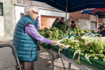 Mujer mayor comprado verduras en un mercado callejero y hablando por el teléfono móvil. - obrazy, fototapety, plakaty