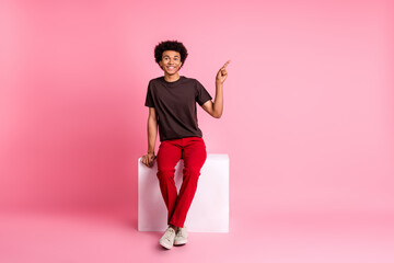 Full body photo of friendly guy sitting platform cube directing finger empty space great deal proposition isolated on pink color background
