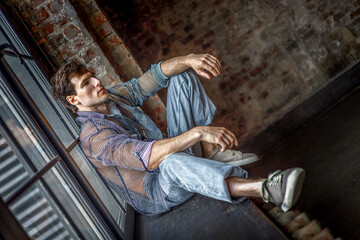 A modern young man with a stylish hairstyle sits on a windowsill near a window on a dark background.