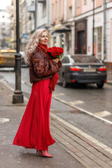 Obraz premium An elegant blonde in a red evening dress and fur coat is standing on the streets of the city, waiting for a taxi. Spring concept, international women's day. March 8