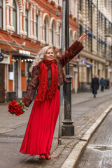 An elegant blonde in a red evening dress and fur coat is standing on the streets of the city, waiting for a taxi. Spring concept, international women's day. March 8