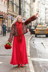 Obraz premium An elegant blonde in a red evening dress and fur coat is standing on the streets of the city, waiting for a taxi. Spring concept, international women's day. March 8