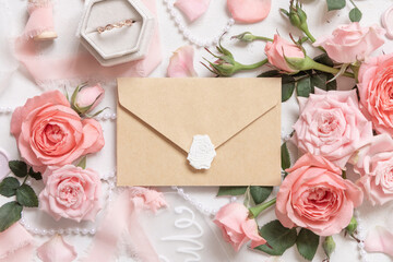 Beige sealed envelope near pink roses, engagement ring and ribbons top view, wedding mockup