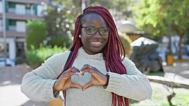 A cheerful woman with braids making a heart shape with her hands in a sunny park