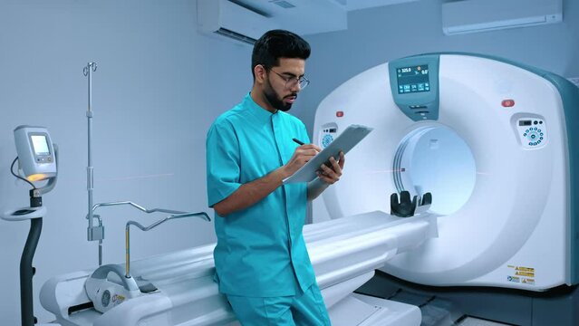 Indian tomographer making notes with pen. Serious doctor at background of CT scanner. MRI specialist preparing for CT scanning. Modern equipped cabinet for MRI examination. MRI capsule.