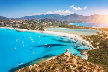 Aerial view of white sandy beach, old tower on the hill, sea bay, mountains at sunset in summer. Porto Giunco in Sardinia, Italy. Top view of blue sea with clear water, white sand, mountains