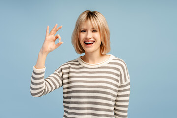 Happy blonde haired lady wearing braces gesturing okay while looking at the camera