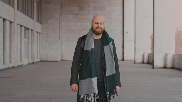 Gimbal footage of cheerful confident bearded man in coat and scarf walking near white building in city.