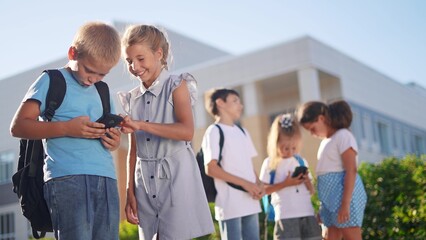 children group school children look at the smartphone video. school learning kid concept. group of kids with backpacks playing smartphone near the school building sunlight outdoors