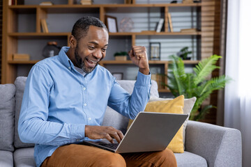 Ecstatic African American man raising fist in victory while using laptop on couch, feeling excited and happy.