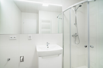 Fototapeta na wymiar Bright white small bathroom with sink and shower. The bathroom has a large mirror with led tape, several electrical sockets, heating ladder and water with waste for the washing machine.