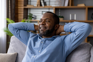 A content African American male sitting on a sofa at home, hands behind head, with a look of...