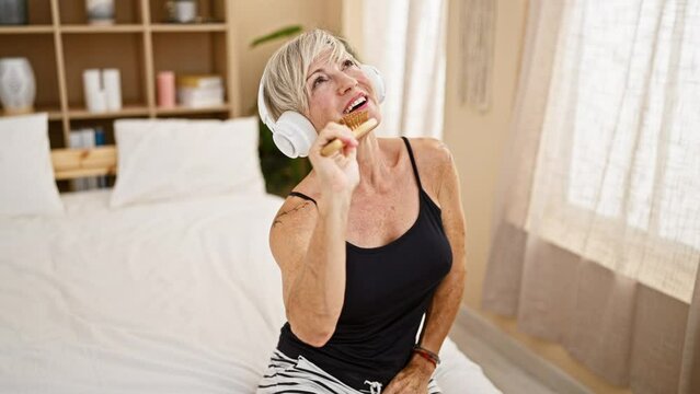 Mature woman with headphones enjoys music while holding a hairbrush and singing on a bed in a bedroom setting