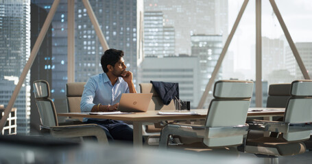 Portrait of a Young Indian Account Manager Working on a Laptop Computer in a Modern Corporate Office. Businessman Dealing with Financial Reports, Preparing a Growth Plan for the Company - obrazy, fototapety, plakaty