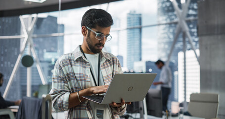 Indian Male Business Analyst Standing with Laptop Computer in a Modern Office. Successful Man Assessing Technological Risks And Developing Strategies For Company Growth and Expansion