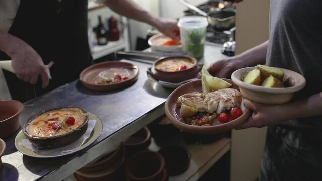Inside the restaurant kitchen. Closeup view of a waitress holding a fish filet and potatoes garnish dish, waiting for the chef to finish crab pies served in clay bowls. 