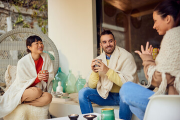 A group of three friends sitting outdoors, covered in a blanket, drinking coffee and talking to each other. - 753136007