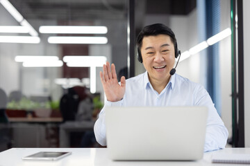 Asian customer support agent with headset smiling and waving at the camera during a video call in a...