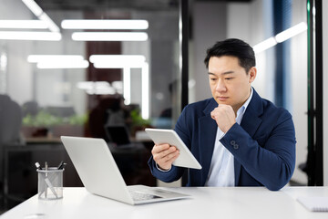 A professional businessman thoughtfully using a digital tablet, with a laptop open in his well-lit office space.