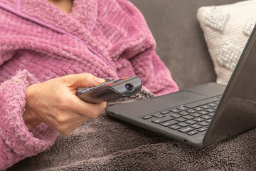 Woman sitting on sofa at home with TV remote control and laptop on her lap
