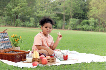 Happy smiling African little girl with curly hair enjoys a picnic in summer park. Kid eating bread...