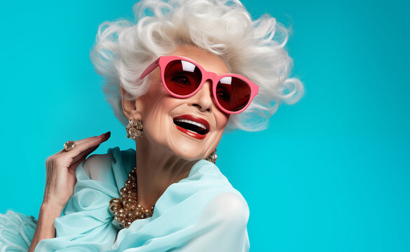 Fancy eldery late 60s grey heir woman funy pink glasses happy laughing on the blue wall background. Elderly happy people and calm finance positive retirement concept image.