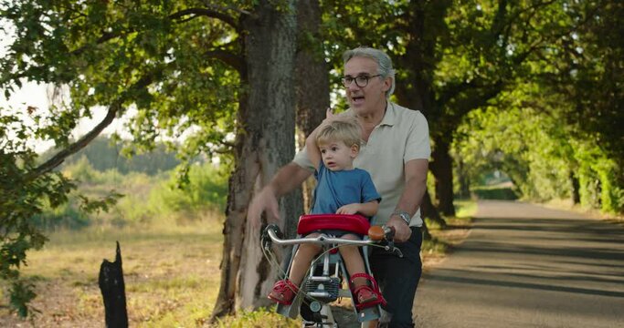 At sunset, a young grandfather takes his grandson on his bike along a road surrounded by nature and they have fun together