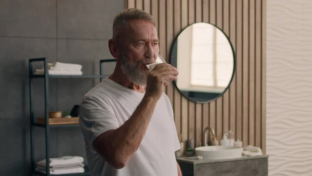 Old Caucasian man senior mature retired male in bath bathroom morning drinking still water drink holding glass refreshing healthy habit aqua balance h2o refresh thirst health care elderly generation