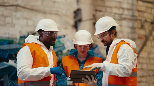 trio of happy successful workers in white uniform and orange vest reviewing their plans on a tablet in a large hall. A man with Black skin a brunette girl and a guy with a beard are talking and