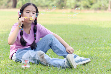 Happy cute Asian girl with pigtails blowing soap bubbles while sitting on green grass in nature...
