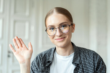  Smiling young woman with glasses greeting with a wave in a bright, airy room.