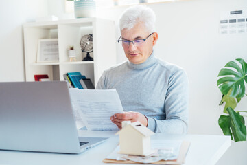 Middle aged man working on laptop with house model money box and docs on table. Housing cost and payments. Real estate, property investment. Home mortgage loan rate. Saving money for retirement.