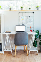 Modern cozy light workplace - white desk with laptop mockup empty screen, grid mood board with pinned notes, shelves with docs and green monstera plant at work space in home office room interior