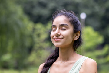 Close-up photo of a smiling and relaxed young Indian woman standing outside in a park and resting...