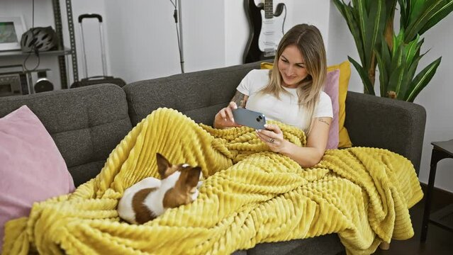 A smiling young woman takes photos of her cute dog while relaxing on a yellow blanket on a couch at home.